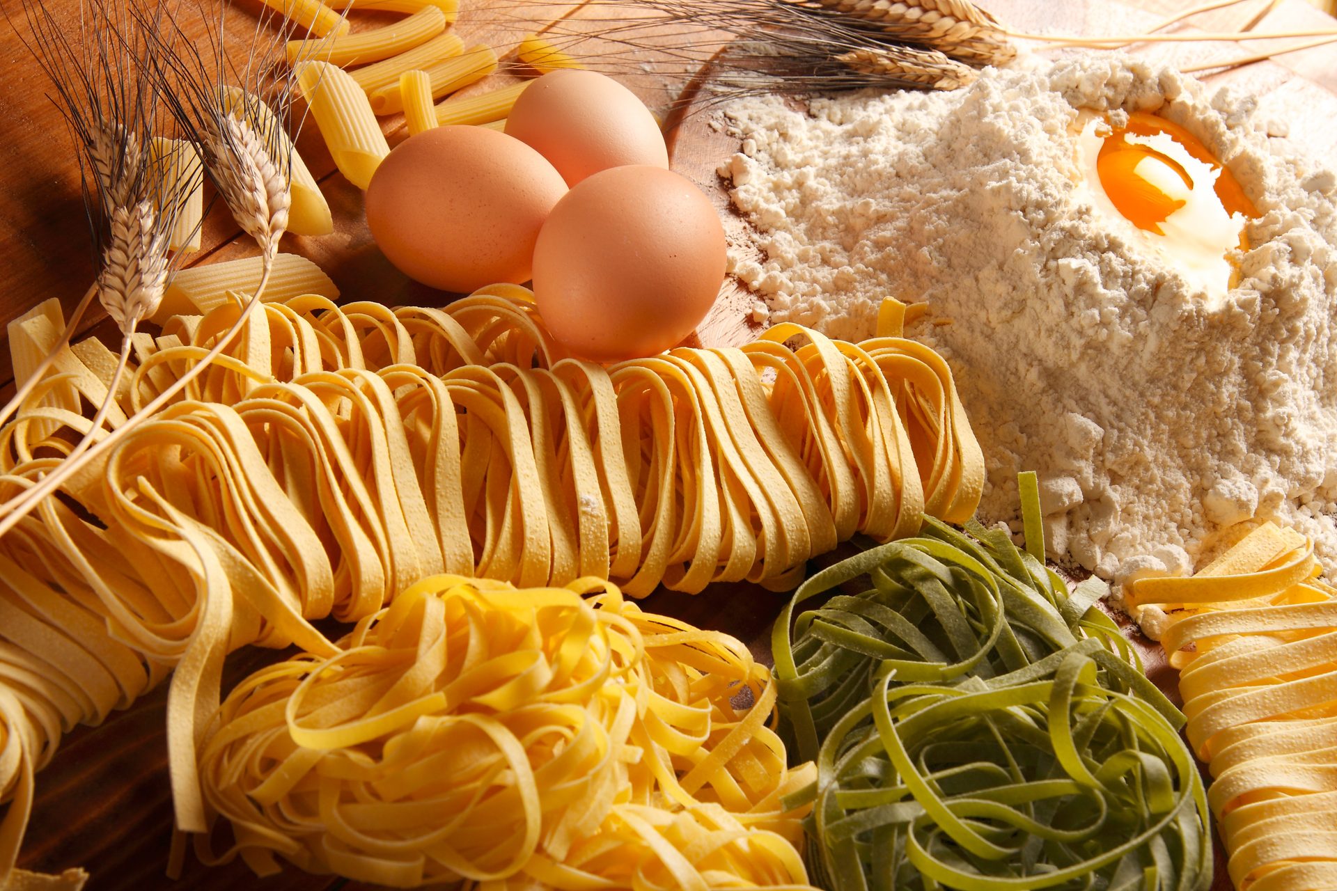 Nidi di tagliatelle gialle e verdi, penne, tre uova, spighe di grano e farina con tuorlo su un tavolo di legno